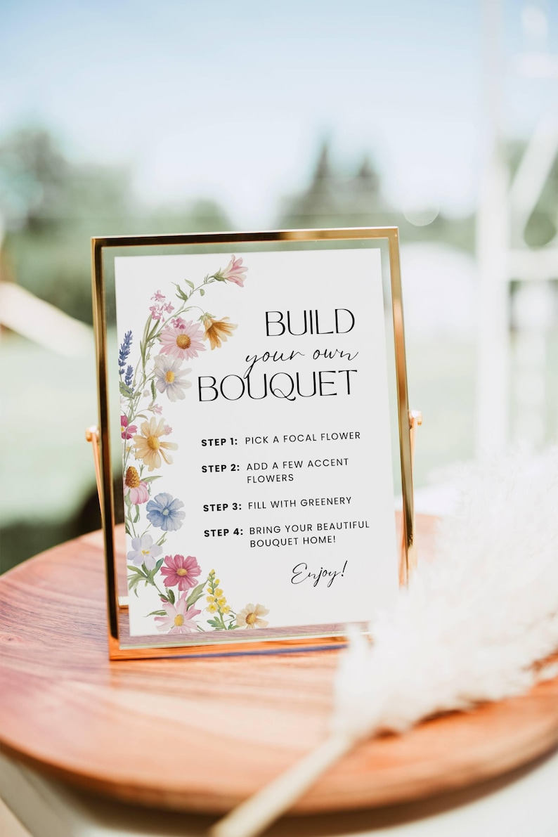 May include: A gold-framed sign with the text "Build Your Own Bouquet" and instructions. The sign is decorated with watercolour flowers in pink, yellow, and blue. The sign is on a wooden table.