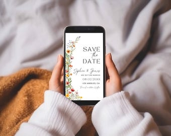 Op de afbeelding: Een smartphone met een digitale Save the Date-huwelijksaankondiging. Het ontwerp bevat een verticale schikking van kleurrijke aquarel-wilde bloemen aan de linkerkant en zwarte tekst met Save the Date, Sophia & James, 08.02.20XX, Los Angeles, CA.