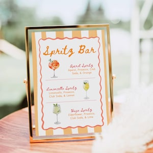 Może przedstawiać: Złota ramka z napisem "Spritz Bar" w pomarańczowym kolorze. Znak wymienia trzy koktajle spritz: Aperol Spritz, Limoncello Spritz i Hugo Spritz, z ich składnikami. Tło to rozmyta scena na zewnątrz.