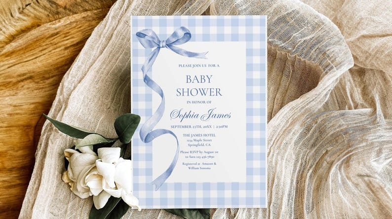 May include: A baby shower invitation with a blue gingham border and a watercolor bow and ribbon. The invitation text reads "Baby Shower" and includes the honoree's name, date, time, and location. White flowers and greenery are in the lower left corner.