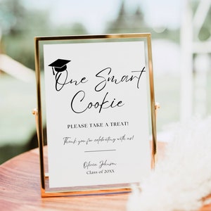 May include: A gold-framed sign with the text "One Smart Cookie" and a graduation cap icon. The sign also includes the text "Please take a treat! Thank you for celebrating with us! Olivia Johnson Class of 20XX".