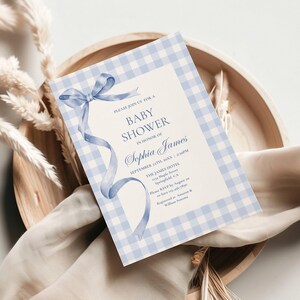 May include: A baby shower invitation with a light blue gingham border and a watercolor ribbon design. The invitation includes the text "Baby Shower" and the name "Sophia James". The invitation is on a wooden tray with dried plants.