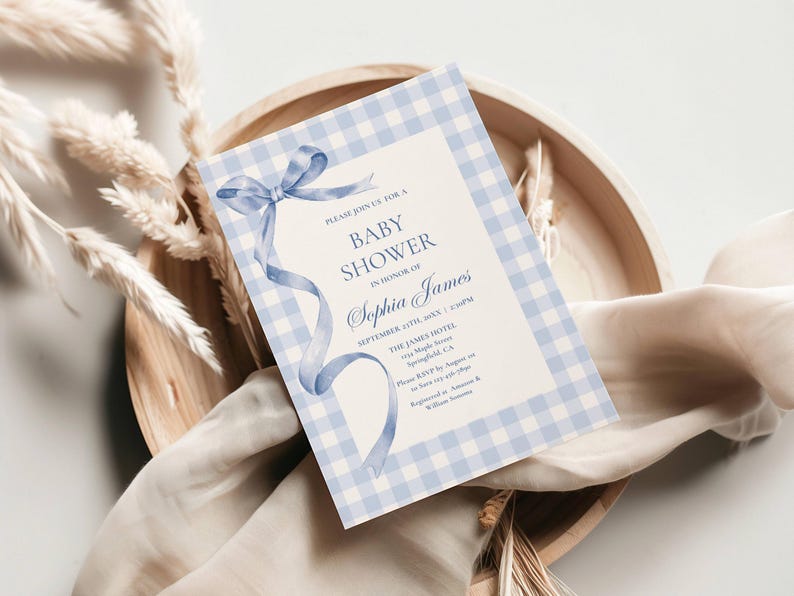 May include: A baby shower invitation with a light blue gingham border and a watercolor ribbon design. The invitation includes the text "Baby Shower" and the name "Sophia James". The invitation is on a wooden tray with dried plants.