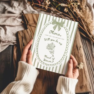 May include: A green and white striped invitation card with the text "Olivia, I can't say I do without you! Will you be my Maid of Honor?" is held by a person. The card features a bow and floral bouquet illustration.