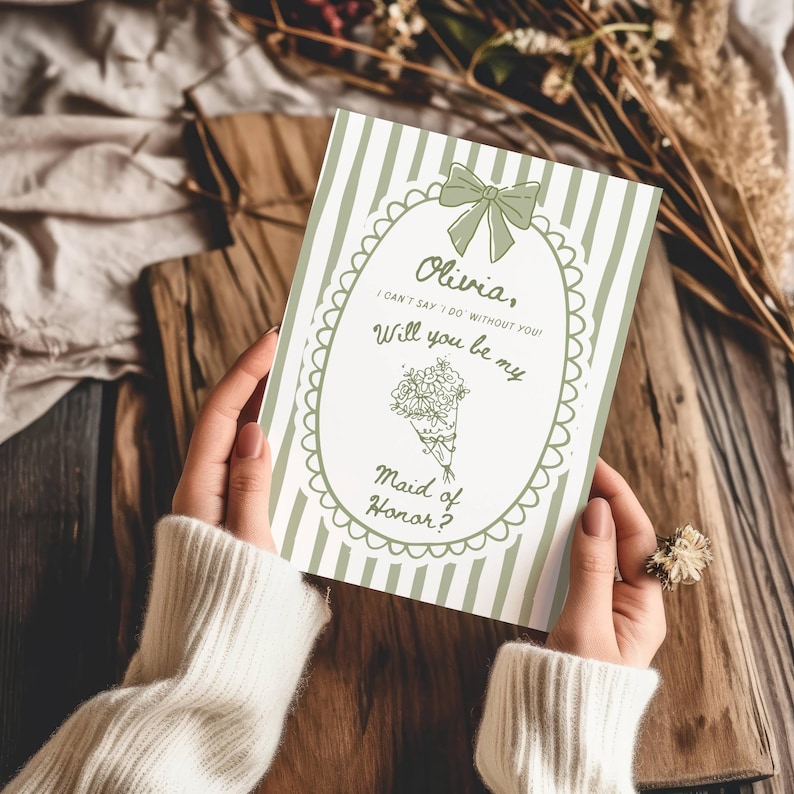 May include: A green and white striped invitation card with the text "Olivia, I can't say I do without you! Will you be my Maid of Honor?" is held by a person. The card features a bow and floral bouquet illustration.