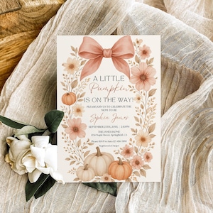 May include: A baby shower invitation with a floral and pumpkin theme. The card features a peach bow, flowers, and pumpkins. Text reads "A Little Pumpkin is on the Way!" and includes event details. The invitation is set on a beige fabric.