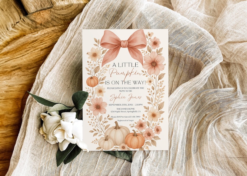 May include: A baby shower invitation with a floral and pumpkin theme. The card features a peach bow, flowers, and pumpkins. Text reads "A Little Pumpkin is on the Way!" and includes event details. The invitation is set on a beige fabric.