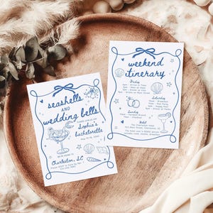 May include: Two white rectangular cards with blue ink illustrations and text. One card reads seashells and wedding bells for Sophia's bachelorette in Charleston, SC. The other card is a weekend itinerary with a schedule of events and icons.
