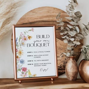 May include: A framed sign with the text "Build Your Own Bouquet" and instructions, surrounded by floral illustrations. The sign is displayed on a wooden surface with decorative vases and dried plants, creating a floral arrangement theme.