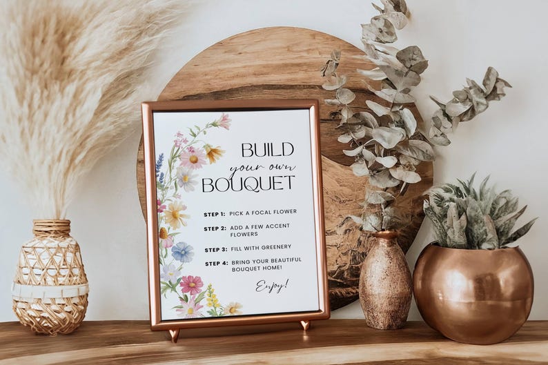 May include: A framed sign with the text "Build Your Own Bouquet" and instructions, surrounded by floral illustrations. The sign is displayed on a wooden surface with decorative vases and dried plants, creating a floral arrangement theme.