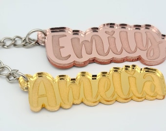 Puede incluir: Dos llaveros de color dorado con los nombres "Emily" y "Amelia" escritos en letra cursiva. Los llaveros están superpuestos y tienen un acabado espejado.