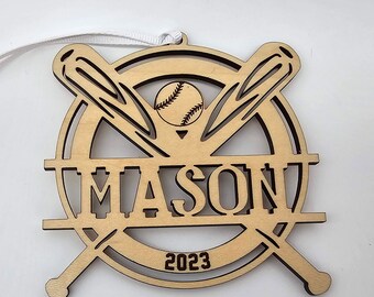 Puede incluir: Adorno de madera de béisbol con un diseño de béisbol y bates. El nombre "MASON" está escrito en el centro del adorno. El año "2023" está escrito en la parte inferior del adorno.