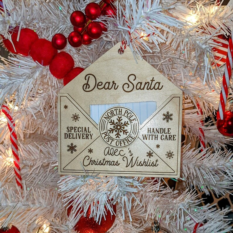 Puede incluir: Un adorno navide&ntilde;o de madera con forma de sobre con las palabras "Querido Santa" en la parte superior. El adorno tiene un espacio para escribir una carta a Santa e incluye el texto "Polo Norte Oficina de Correos", "Entrega Especial", "Manejar con cuidado", y "Lista de deseos de Navidad de Alec".