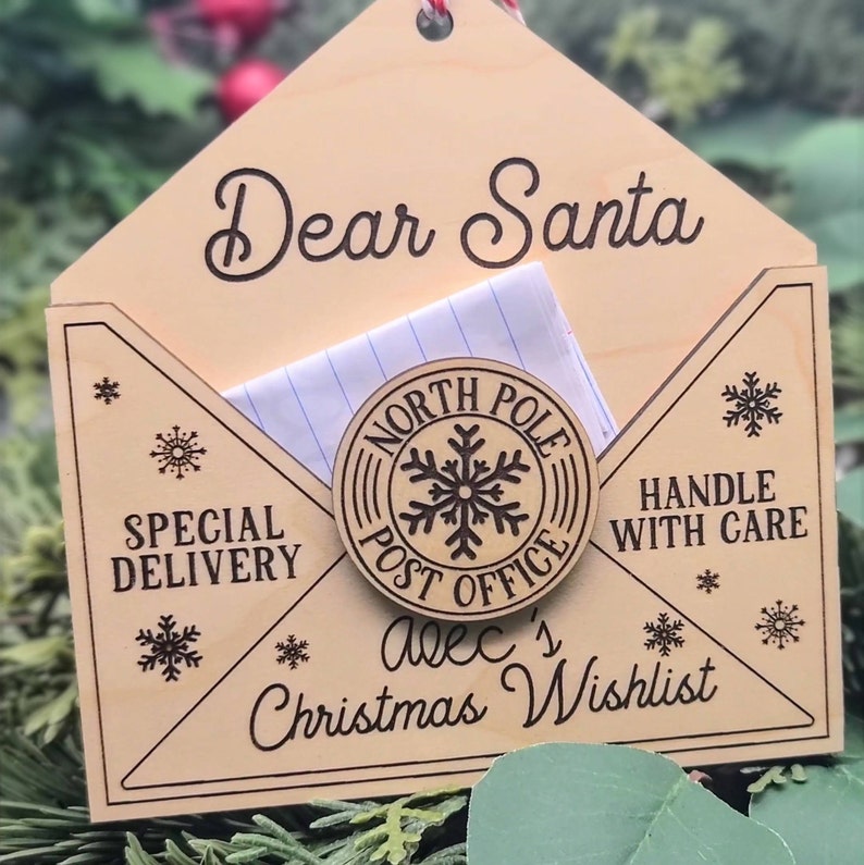 Puede incluir: Un adorno navide&ntilde;o de madera con forma de sobre con una ranura para una carta. El adorno est&aacute; decorado con las palabras "Querido Santa", "Entrega especial", "Oficina de correos del Polo Norte", "Manejar con cuidado" y "Lista de deseos de Navidad de Alec".