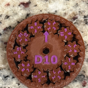 May include: A brown plastic D10 dice with a purple inscription. The dice is made up of ten smaller gears, each with a number from 1 to 10 printed on it. The gears are arranged in a circle around a central gear, which is labelled "D10".