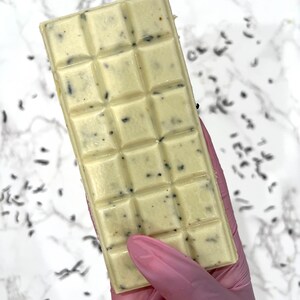 May include: A white chocolate bar with dark flecks throughout. The bar is divided into squares and is being held by a gloved hand.