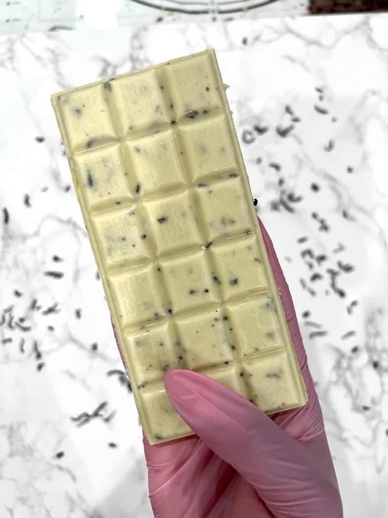 May include: A white chocolate bar with dark flecks throughout. The bar is divided into squares and is being held by a gloved hand.