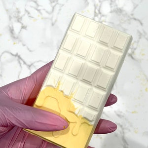May include: A white chocolate bar with a yellow chocolate drizzle. The chocolate bar is held in a pink latex glove.