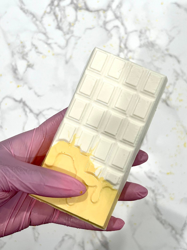 May include: A white chocolate bar with a yellow chocolate drizzle. The chocolate bar is held in a pink latex glove.