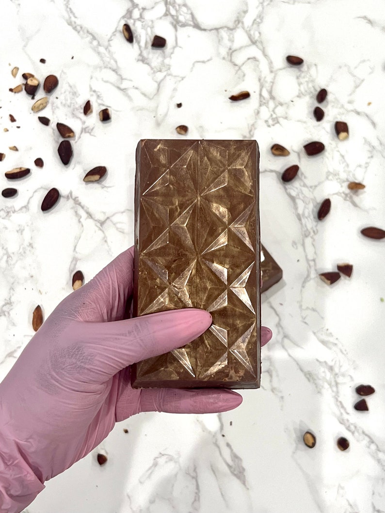 May include: A single bar of dark chocolate with a geometric pattern on the surface. The chocolate bar is being held by a gloved hand.