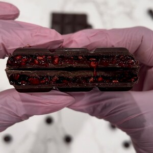 May include: A close-up of a dark chocolate bar with a raspberry filling. The bar is broken in half, revealing the red raspberry filling.