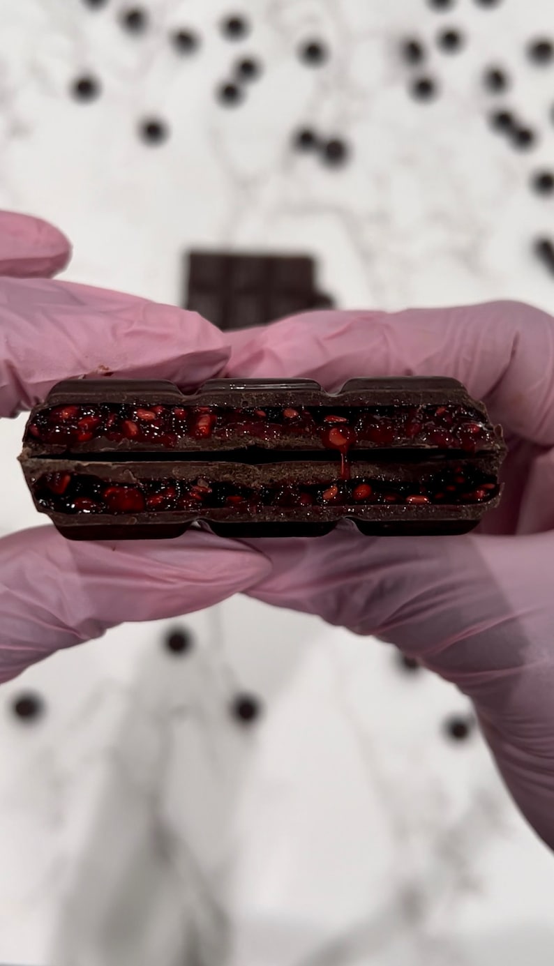 May include: A close-up of a dark chocolate bar with a raspberry filling. The bar is broken in half, revealing the red raspberry filling.