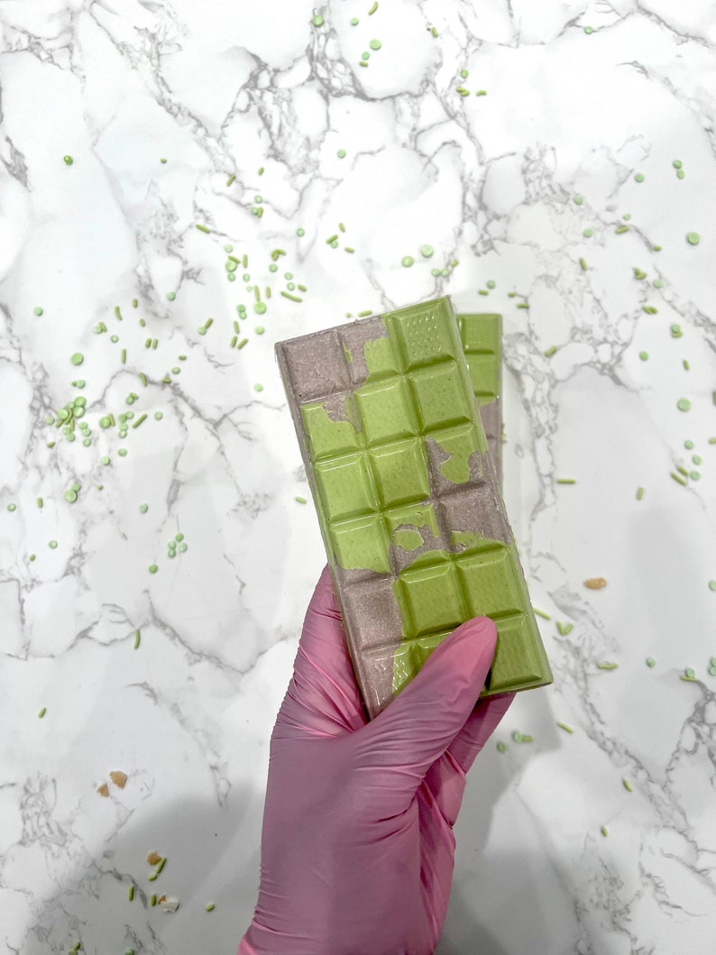 May include: Two green and gray chocolate bars with a marbled design. The bars are held in a pink latex glove against a white marble background.