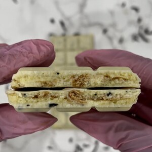 May include: A white chocolate bar with a layer of cookie crumbles in the middle. The bar is broken in half, revealing the cookie crumbles.
