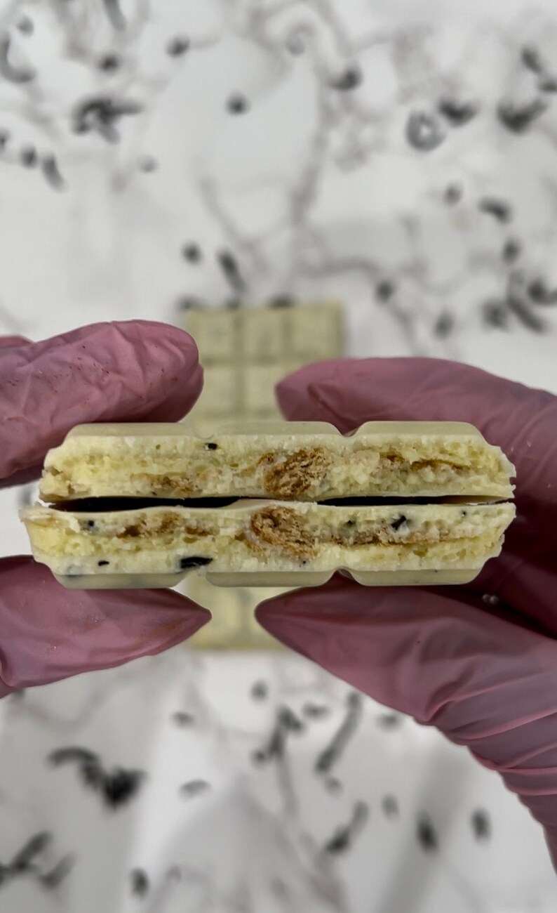 May include: A white chocolate bar with a layer of cookie crumbles in the middle. The bar is broken in half, revealing the cookie crumbles.