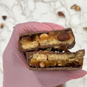 May include: A close-up of a chocolate bar broken in half, revealing a layer of caramel, almonds, and graham crackers.