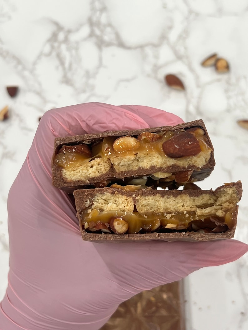 May include: A close-up of a chocolate bar broken in half, revealing a layer of caramel, almonds, and graham crackers.
