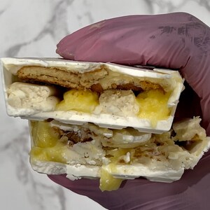 May include: A white chocolate bar with a layer of lemon curd and a layer of cookie crumbs. The bar is broken in half, revealing the layers inside.