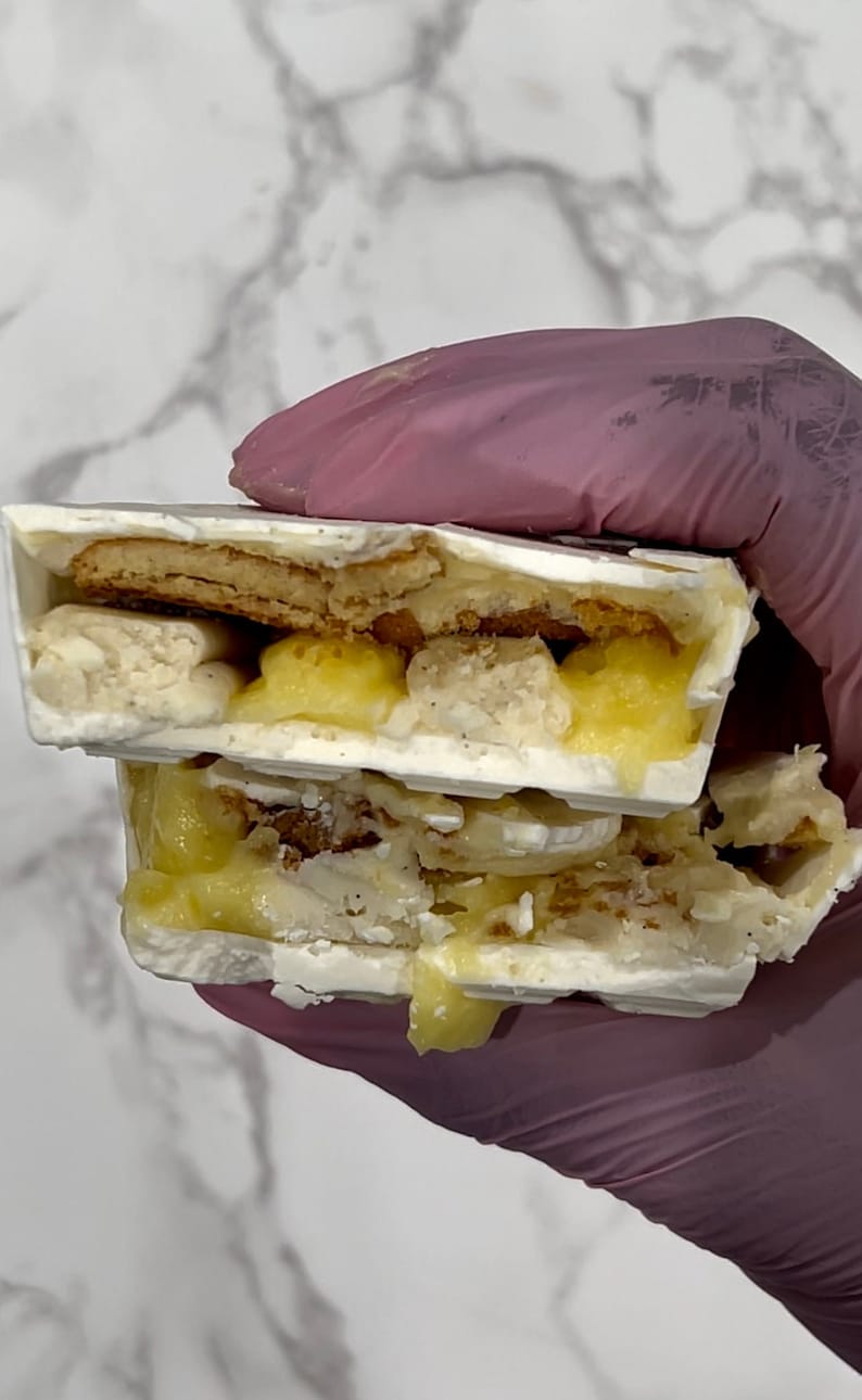 May include: A white chocolate bar with a layer of lemon curd and a layer of cookie crumbs. The bar is broken in half, revealing the layers inside.