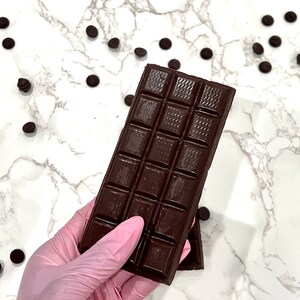 May include: A dark chocolate bar with squares broken into smaller pieces, held in a pink latex glove. The chocolate bar is on a white marble surface with scattered dark chocolate chips.