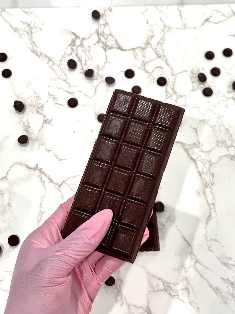 May include: A dark chocolate bar with squares broken into smaller pieces, held in a pink latex glove. The chocolate bar is on a white marble surface with scattered dark chocolate chips.
