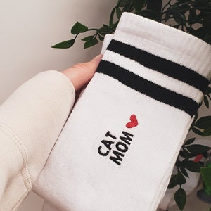 Könnte beinhalten: Weiße Socken mit schwarzen Streifen und dem gestickten Text "Cat Mom" in Schwarz mit einem roten Herzen.