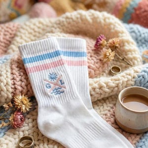 May include: White socks with blue and pink stripes, featuring an embroidered design of a sun, waves, and crossed lines with the letters "G" and "L". The socks are placed on a colourful knitted blanket, with a cup of tea.