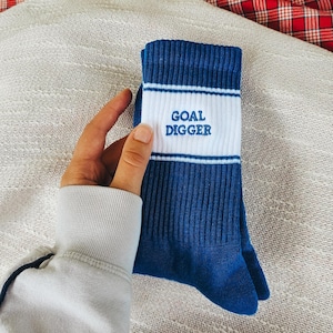 Könnte beinhalten: Ein Paar blaue Socken mit einem weißen Band mit dem Text "GOAL DIGGER" in fetter Schrift. Die Socken haben weiße Streifen über und unter dem Text. Die Socken sind aus einem weichen Material.
