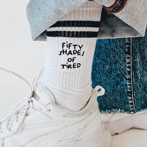 Könnte beinhalten: Weiße Socken mit schwarzen Streifen und dem gestickten Text "Fifty Shades of Tired". Die Socken werden mit weißen Turnschuhen getragen.