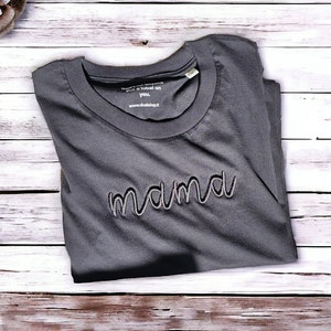 Könnte beinhalten: Ein dunkelgraues T-Shirt mit dem gestickten Wort "mama" in Schreibschrift. Das Shirt ist gefaltet und liegt auf einer hellen Holzmaserung. Der Text "put a label on you. www.duskshop.it" ist auf dem Kragen aufgedruckt.