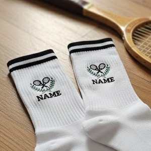 Può includere: Calzini sportivi bianchi con strisce nere, con un design di racchetta e pallina da tennis con la scritta "NAME". Un'antica racchetta da tennis in legno e una pallina da tennis sono anch'esse nell'immagine.