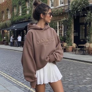 Puede incluir: Una mujer con una sudadera marrón con "SALTY" bordado en la parte delantera, una falda plisada blanca, mocasines negros y un bolso negro. Está de pie en una calle adoquinada frente a un edificio con plantas trepadoras.
