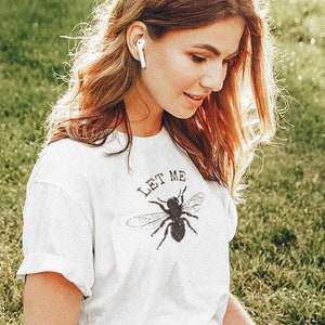 May include: A white t-shirt with the text "LET ME" above a black bee graphic. The person is wearing gray pants and white sneakers while sitting on the grass with a laptop and a coffee cup.