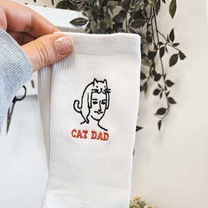 Könnte beinhalten: Weiße Socken mit einer schwarzen und weißen Stickerei, die ein auf dem Kopf einer Person sitzendes Katzenmotiv zeigt. Der Text "CAT DAD" ist in Rot unter der Illustration gestickt.