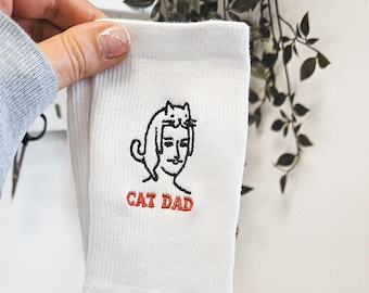 May include: White socks with a black and white embroidered illustration of a cat sitting on a person's head. The text "CAT DAD" is embroidered in red below the illustration.