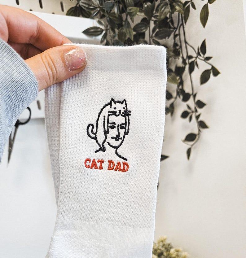 Peut inclure: Chaussettes blanches avec une illustration brod&eacute;e en noir et blanc d'un chat assis sur la t&ecirc;te d'une personne. Le texte "CAT DAD" est brod&eacute; en rouge sous l'illustration.