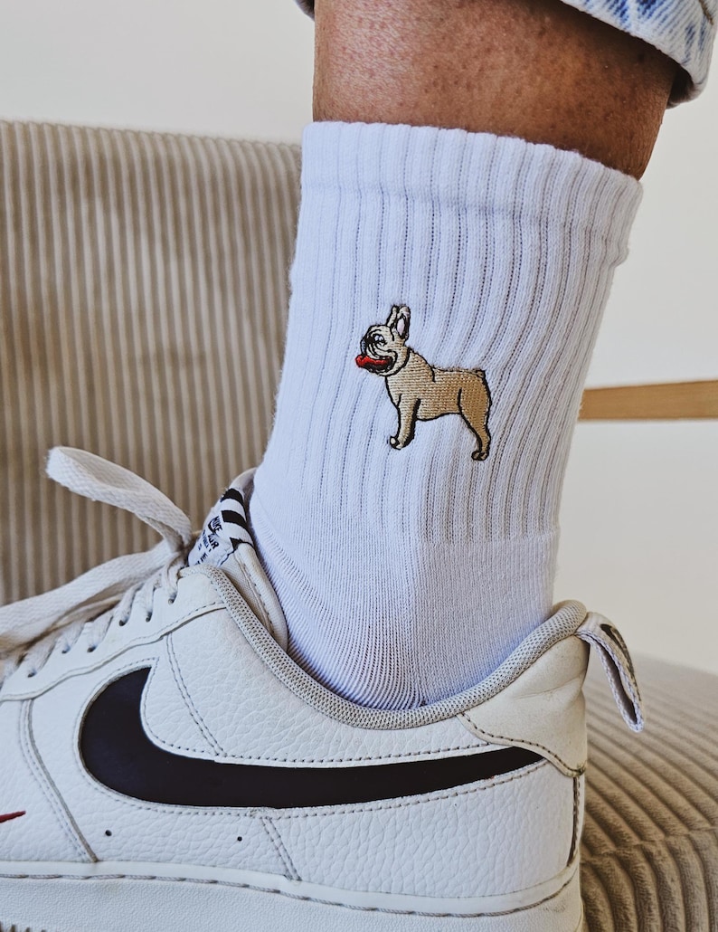 May include: White socks with a small embroidered image of a French bulldog wearing a red collar. The socks are being worn with white trainers with a black swoosh.