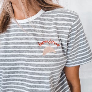 Puede incluir: Camiseta a rayas grises y blancas con un "bonjour" bordado en rojo y un gr&aacute;fico de croissant.