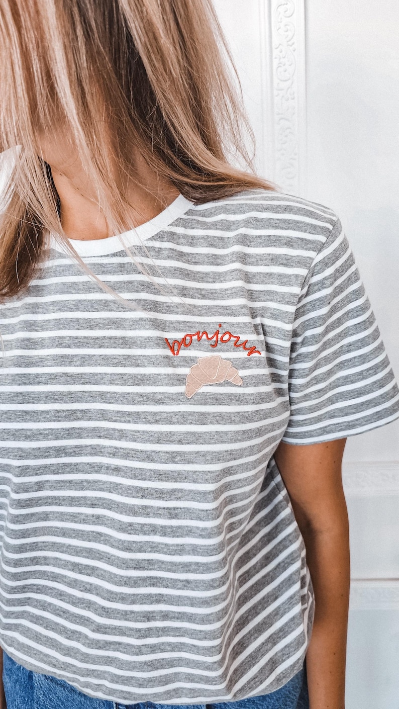 Puede incluir: Camiseta a rayas grises y blancas con un "bonjour" bordado en rojo y un gr&aacute;fico de croissant.