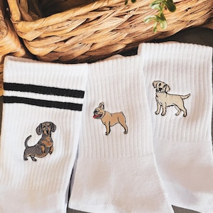 May include: Three pairs of white crew socks with embroidered dog breeds. One pair features a dachshund, another a French bulldog, and the last a Labrador retriever.
